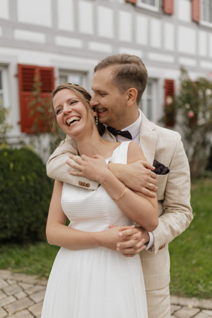 Lachendes Brautpaar vor einem Fachwerkhaus, umarmt sich auf einem Weingut in Franken.