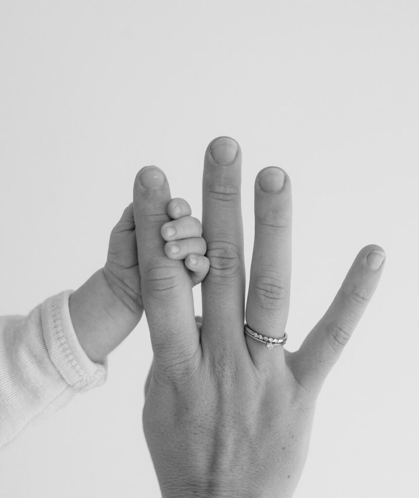 Detailbild in schwarzweiß von der Hand eines Neugeborenem und dessen Mutter mit Verlobungs- und Ehering.